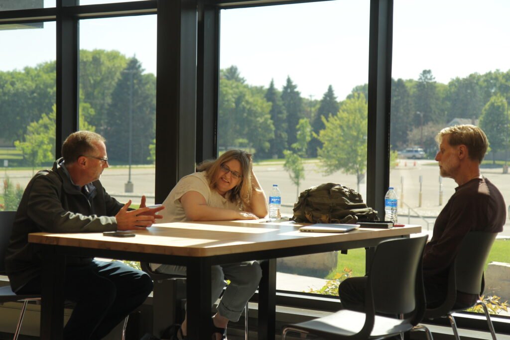 Three people sit at a table in front of a floor-to-ceiling window. Sensory-sensitive learners can achieve more through partnership, facilitation of agency, and strengths-based learning strategy.