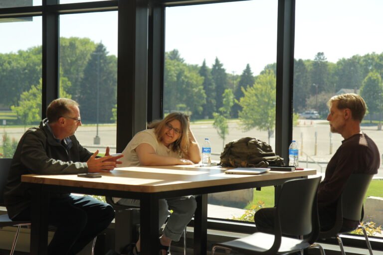Three people sit at a table in front of a floor-to-ceiling window. Sensory-sensitive learners can achieve more through partnership, facilitation of agency, and strengths-based learning strategy.
